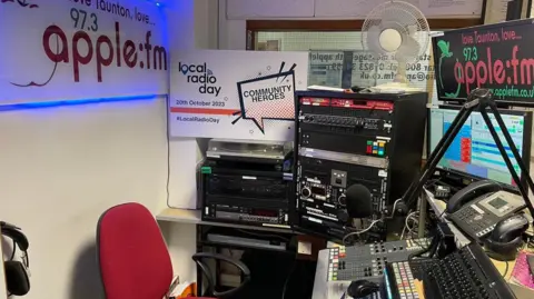 A small radio studio with lots of signs that say Apple FM. There's a window in the background and in the foreground there's radio equipment, a red chair and a microphone.