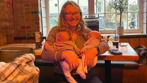 Jess Nicholson Jess holds her twin girls. She appears to be in a pub or restaurant. She has blonde, curly hair and glasses. 