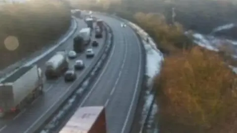 An image from a roadside camera of queuing traffic on the eastbound carriageway of the A14. The road, which is going up a hill and curving around to the left at the top of the picture, has four lanes. The queue is on the two lanes on the left. The other two lanes are clear. There is snow on the tree-lined verges. 