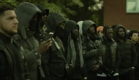 Kay S. Ubhi/Miracle Media Men stand in a line, looking at something unseen. They all have serious expressions on their faces and are all dressed in black jackets, some with teir hoods up and their faces partially covered.