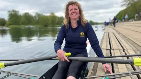 Kate Lindgren wearing a blue fleece sitting in a rowing boat holding her oars. Her left hand is leaning on wooden decking, and in the left of the photo and in the background is the river and some trees on the bank.