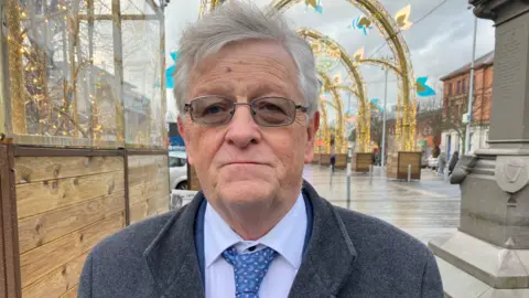 BBC An older man with white hair and glasses standing outside in a pedestrianized part of a town. He is wearing a grey coat over a blue suit, shirt and blue pattern tie. Christmas decorations, mainly lights, can be seen outside along the street.