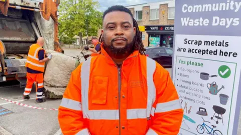 A man in a high-vis jacket stood in front of a bin lorry with a burned mattress being thrown into the back of it. To the right of him is a banner advertising a Community Waste Day with information about scrap metal.