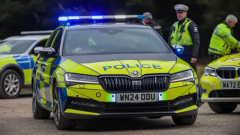 A black, yellow and blue police car with its blue lights on leaves a car park. It is a Skoda and has the liecence plate WN24 0DU. A couple of other police cars are in the car park. Three uniformed officers are standing in the area.