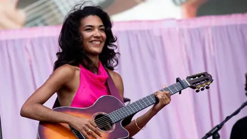 Olivia Dean wears a bright pink dress and plays the guitar, which is on a strap hanging around her. She has long curly black hair and wears gold rings and bracelets