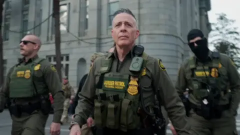 CBS Greg Bovino idzie w mundurze i kamizelce kuloodpornej, po naszej stronie stoją dwie inne osoby w mundurach, jedna z maską. Jest ponuro, w tle widać duży starszy budynek i drzewo