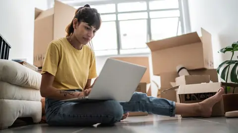 Getty Images Uma pessoa com uma blusa amarela sentada em seu laptop em casa com caixas de mudança atrás dela