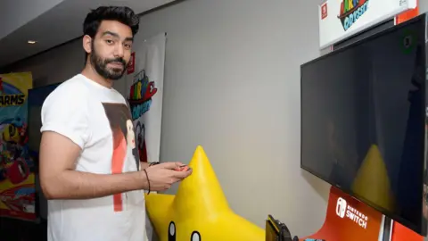 Getty Images Rahul Kohli, wearing a white t-shirt with an image of Princess Leia printed on the front, holds a Nintendo Switch joycon controller as he stands in front of a demo booth for the console. A large statue of Super Mario's invincibility star stands on the floor behind him, and posters for various Nintendo games can be seen in the background.