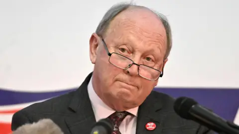 PA A man is standing in front of news microphones. He is wearing a dark suit, white shirt and tie. He has glasses on and is bald with greying hair to the sides.