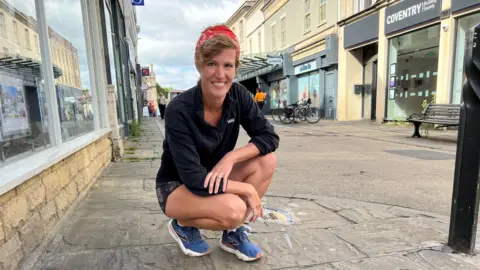 A woman smiles at the camera as she kneels down close to a pavement. She's wearing blue trainers, running shorts, a jacket and a red bandana. Various businesses along the street can be seen behind her.