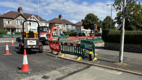 Diggers on a residential road, with green plastic fencing and red and white cones next to them. There are several two-storey, semi-detached houses in the background. The sky is blue with white clouds.