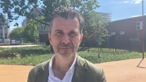 A man with dark and grey hair, wearing a white shirt and green blazer, looks at the camera on a sunny day. Behind him is a sandy colour path, grass and trees, and in the distance are tall buildings. 