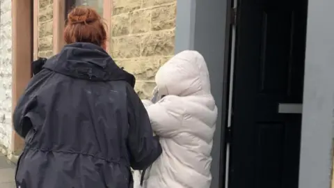 A woman in a cream coat is escorted out of a house by a female police officer in a black coat. The arrested woman has her back to us, and is covering her face with a hood