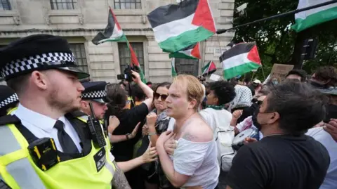 A row of police facing a row of protestors. The protestors, many of which hold Palestinian flags, look angry as they confront the police