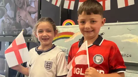 Two younger puplis smile at the camera, they are wearing England football kits and are holding England flags.