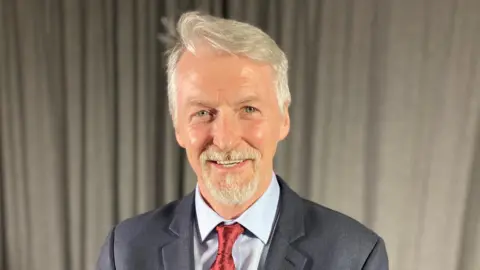 Huw Irranca-Davies, who has grey hair and  a goatee beard smiles at the camera. He is wearing a grey suit and red tie against a pale shirt. Behind him is a beige curtain