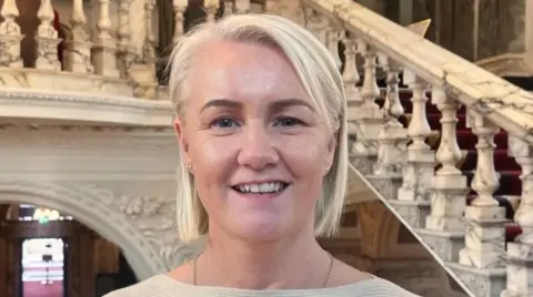 Tracy Kelly standing in Belfast City Hall, she is wearing a cream top and has blonde hair