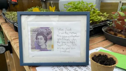 A framed £20 note is on display in a greenhouse, with the message: "Future Jacob, give this money to a young entrepreneur, you'll know when the time is right."