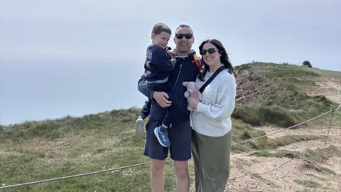 Leanne Rinne/BBC A husband and wife and their young son smiling near the edge of a cliff which is overlooking the sea. The man is holding his son in his arm. 