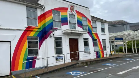 The front of a hospital building with a rainbow painted on it. Disabled parking spaces in front.