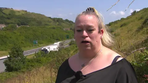 A woman with tied back blonde hair and black top standing on a hill above a motorway.