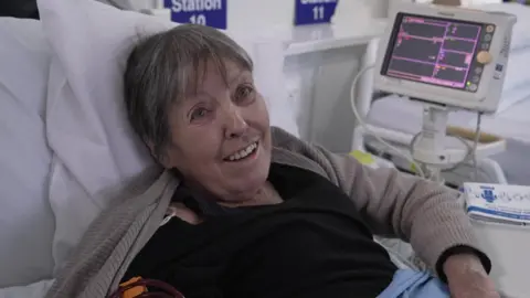 Lewis photographed looking towards the camera from her hospital bed. She is wearing a black tshirt and a beige cardigan. She has short, dark grey hair, and is smiling. She is propped up on white pillows, with a blue hospital blanket. Behind her is a screen, and a blue sign reading 'Station 10' in white letters. 