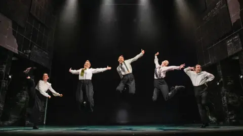 Five actors dressed in white shirts and grey trousers stand on stage while jumping in the air.