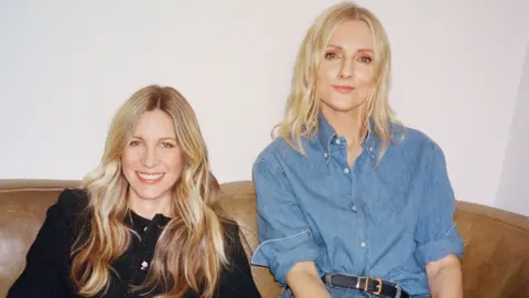 Two women with blonde hair smiling, one wearing a black jacket and the other a blue button up shirt.