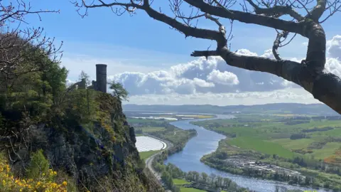 A sunny day from the top of Kinnoull Hill. The sky is blue with a large white cloud. A tower is in the background, surrounded by green plants, gorse bushes and rocks. A river can be seen in the centre of the image. A tree is dark in the top right foreground.