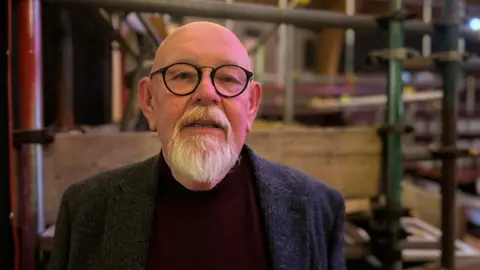 Mark Dornford-May is a man in his sixties who is bald with a white goatee beard. He is wearing a jacket and a brown jumper. He is inside a building and behind him, but out of focus, is a lot of scaffolding. 