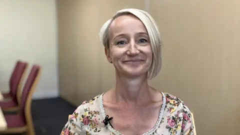 A woman with short blonde hair, which is longer on one side, smiles at the camera. She is wearing a small microphone and has a floral top on. She is smiling. She is sat in a conference room with beige walls and chairs can be seen in the background.