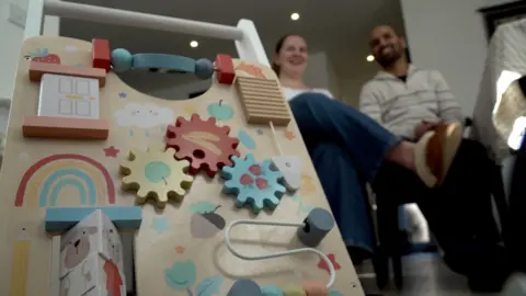 Ben Schofield/BBC A child's wooden toy, with colourful cogs, flaps and moving parts dominates the left of the image. Behind it, and out of focus, are two adults - one female, one male sitting near a dining table. They are casually dressed and are both smiling. 