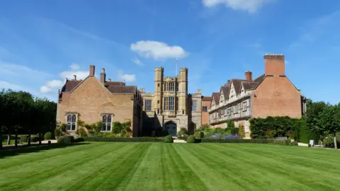 National Trust A huge Tudor-style country house behind vast grounds lined with hedges, trees, and with a footpath around the perimeter. 