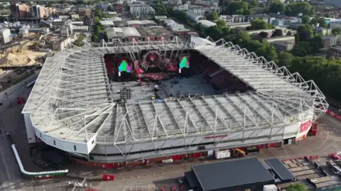 A drone shot looking down on St Mary's Stadium in Southampton. Within the stadium we can just make out a large colourful stage, as preparations for Take That's tour take place.