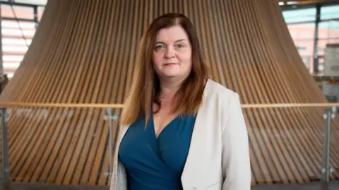 A woman with long brown hair, wearing a blue wrap dress underneath a beige suit jacket stares into the camera in the Oriel in the Senedd.