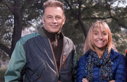 The Winterwatch presenters Chris Packham and Michaela Strachan are standing side by side in a professional picture. Mr Packham has short brown hair and is wearing a brown zip-up fleece, and a blue/ green jacket. Ms Strachan has short blonde hair with a fringe and is wearing a dark blue coat with a patterned scarf. 