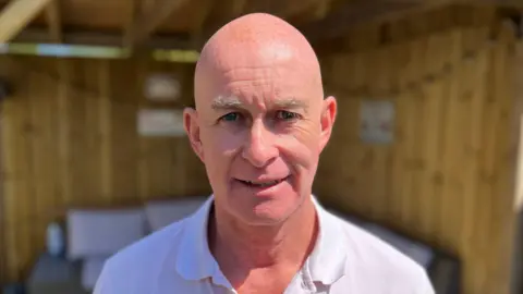 Eamonn Aston smiles into the camera. He's wearing a white polo shirt. He is bald. Behind him is a blurred shed with a visible seating area. 