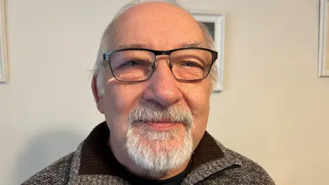 John Devine/BBC Barry Wainwright has very short grey hair on top of his head, he has a short, neatly trimmed grey moustache and beard and black framed glasses.
He is wearing a brownish woollen top with the collar up. Behind him is a magnolia living room wall, with parts of picture frames visible.