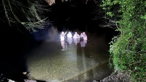 A group of men huddled in a pool. It is still dark and they are wearing swimming shorts. They are all in the water up to their waist and are surrounded by trees. 