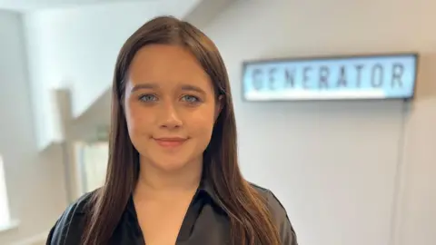 Kaitlin Robson has long brown hair and is smiling at the camera. A sign behind her reads: "Generator".