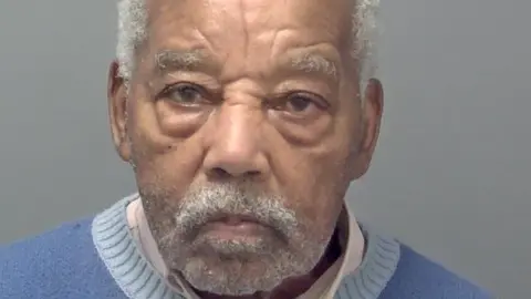 Headley's custody picture. He has white hair, eyebrows and short facial hair, and wears a white collared shirt and a blue jumper. He is looking straight ahead with a serious expression. 