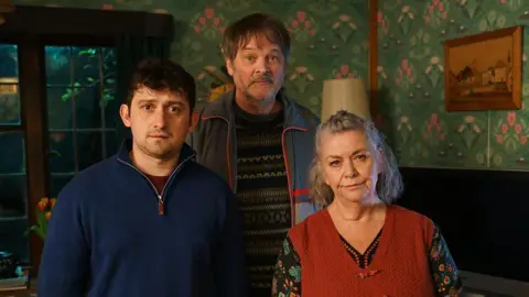 A still from the series showing Craig Roberts, Mark Heap and Dawn French posing as their characters in their dining room. 