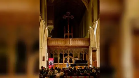 SJE Arts A man performing on a black grand piano in a dimly-lit church. The photo was taken from the back of the church with the audience facing towards the pianist