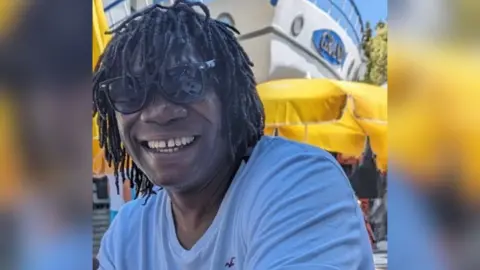 Derek Thomas is smiling at the camera with almost shoulder-length hair in dreadlocks. He is wearing sunglasses and wears a white t-shirt, sitting outside. Behind him are yellow umbrellas and the bow of a boat.