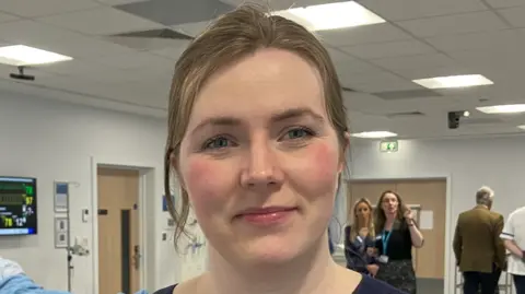 A woman's face dominates the image, which includes walls, doors and other people in the background. The woman has brown hair.  