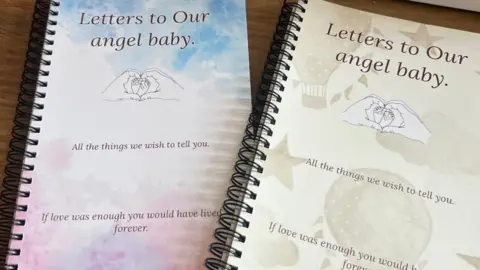 Zoe Wilbourne Two spiral bound journals. They have covers in soft taupe and blues and pink with a drawing of two hands forming a heart shape, round a pair of baby's feet.