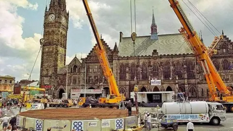 Rochdale Observer Large crane next to milk tankers and huge flat, round cooking surface. A town hall structure is visible in the background.
