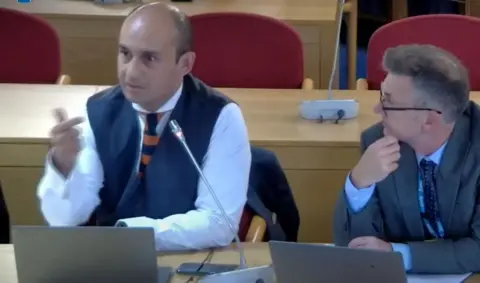 Worcestershire County Council WMAS director Vivek Khashu addressing the council meeting, wearing a shirt, tie and waistcoat, sitting alongside another man in a grey suit