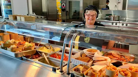BBC A canteen working stands behind trays of cooked dinners, vegetables and fried food.