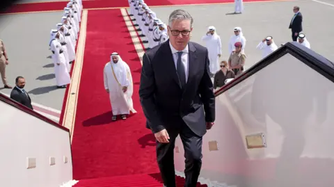 Keir Starmer boarding a plane at the airport in Doha, Qatar. There is a red carpet, lined with men, leading up to the plane